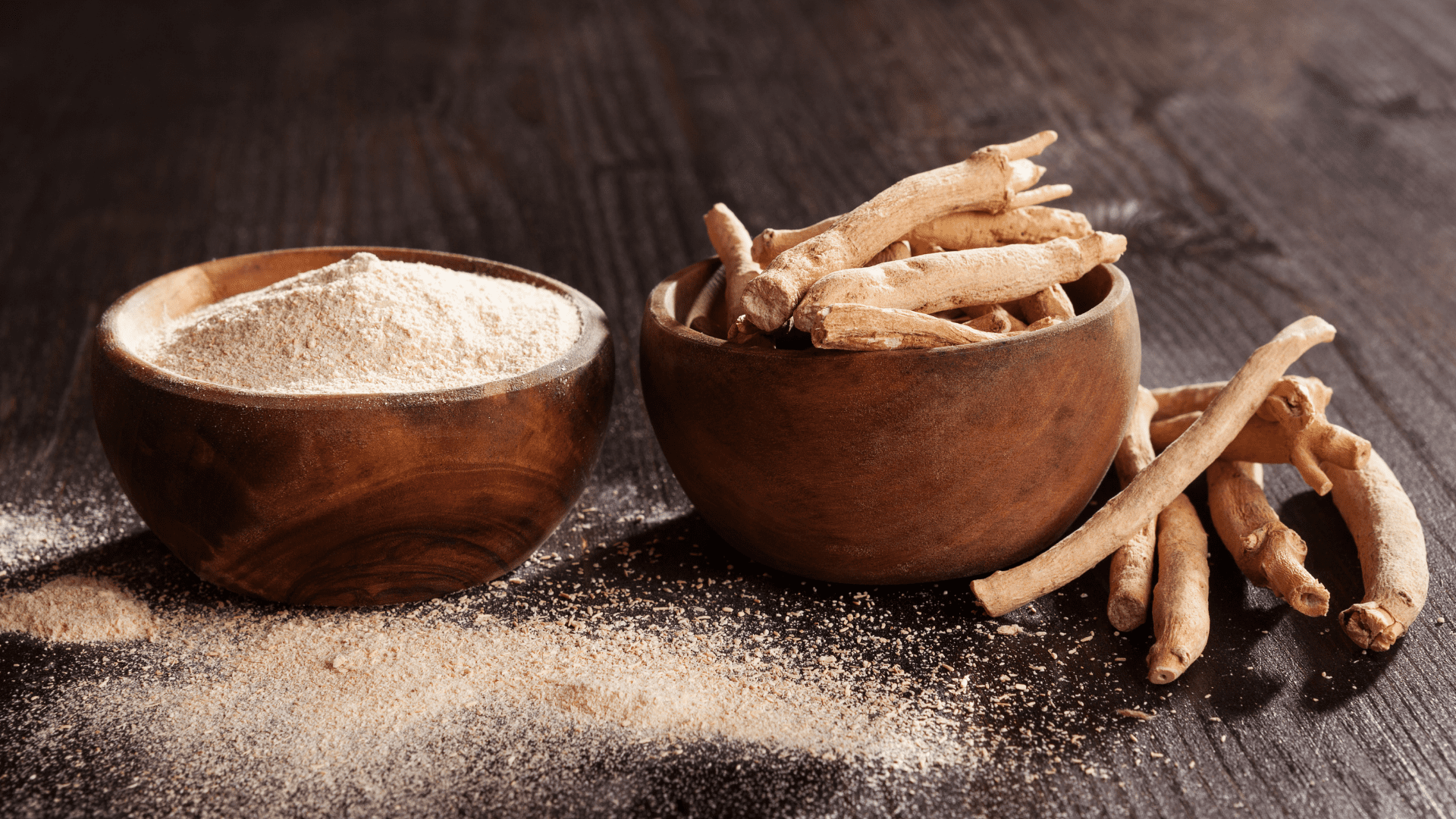 Ashwagandha powder in left wooden bowl, Ashwagandha roots in right wooden bowl, Ashwagandha powder and roots in front and next to the bowls on black wood