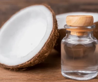 MCT-Öl in kleiner Glasflasche mit Holzdeckel, im Hintergrund Kokosnusshälften auf Holztisch