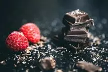 Zwei dunkle Schokoladenstücke gestapelt, daneben Himbeeren, auf einem schwarzen Tisch